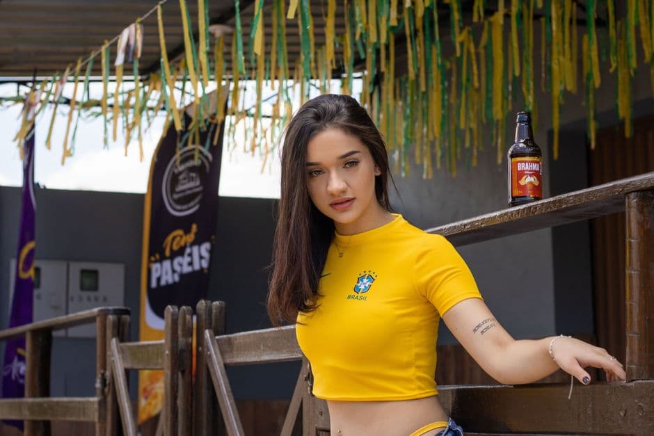 Tattooed woman in a yellow shirt poses at a Brazilian-themed bar with festive decor.