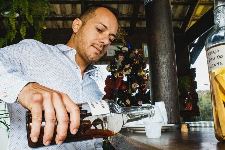 Low angle of young male in classic shirt pouring alcohol drink in shot sitting at wooden counter in summer bar