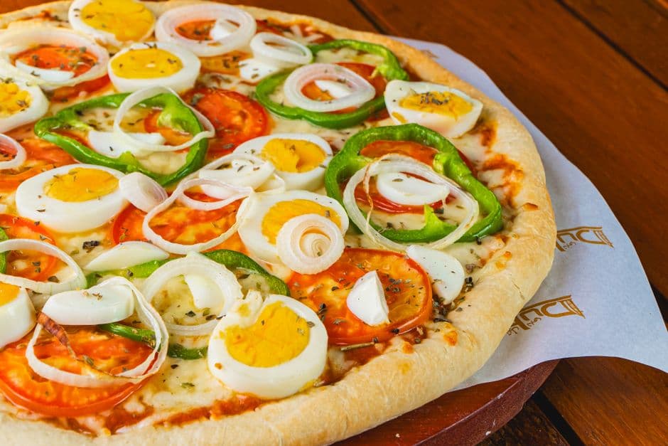 Close-up of a pizza topped with eggs, onions, and colorful vegetables on a wooden table.