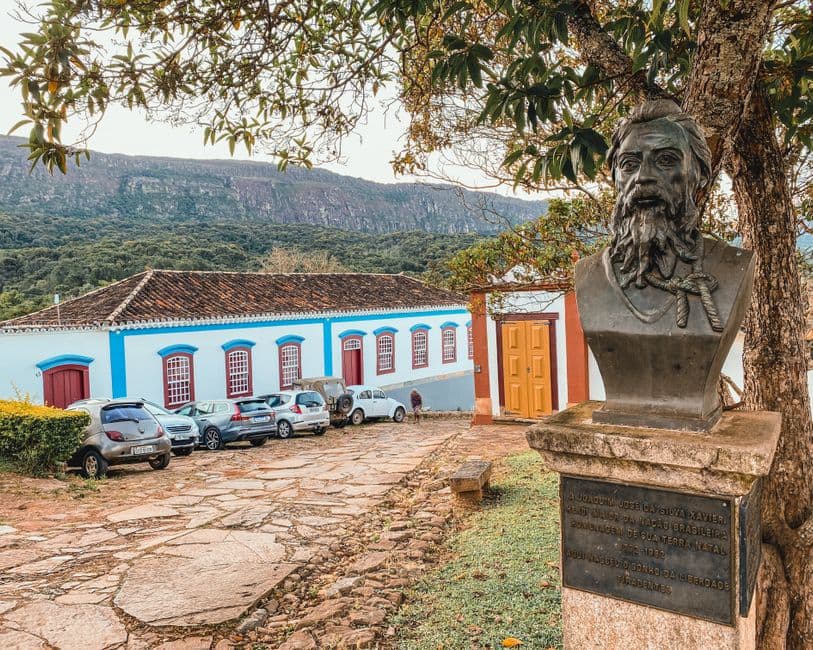A scenic view of Tiradentes, Brazil featuring a historic bust, colonial buildings, and parked cars.