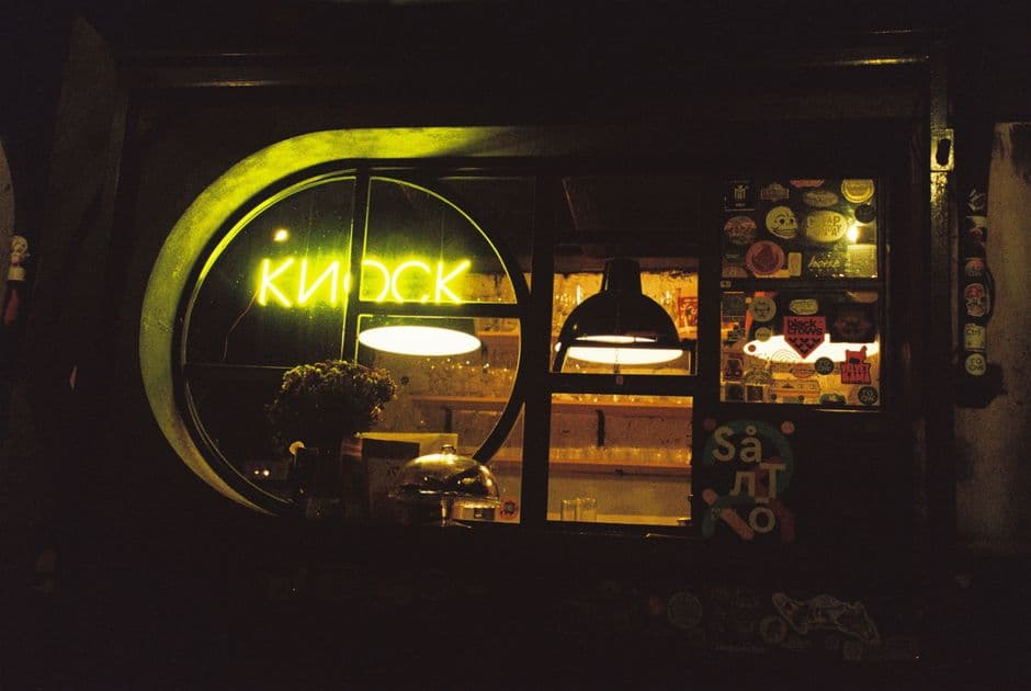 A shop window with a glowing neon sign, warm lighting, and various stickers, creating a cozy atmosphere at night.