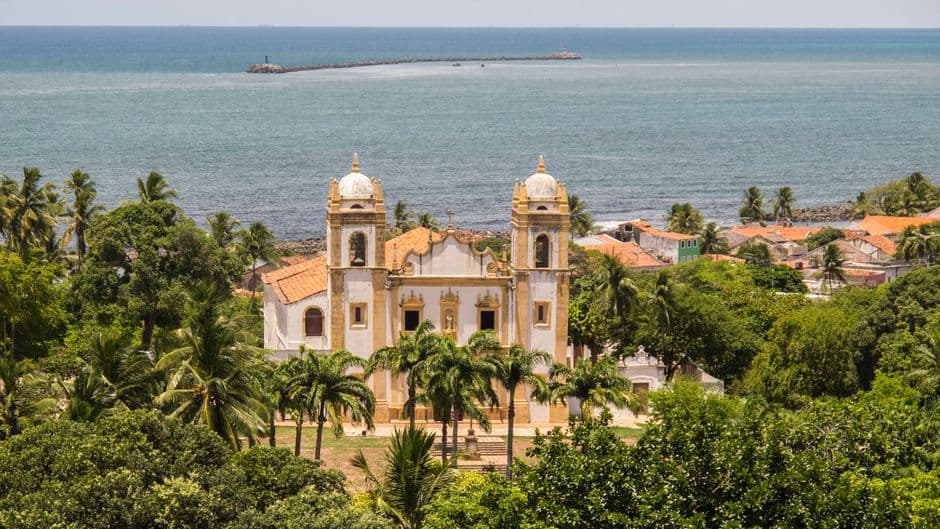 Scenic view of a historic church and lush greenery in Olinda, Brazil with the ocean in the background.