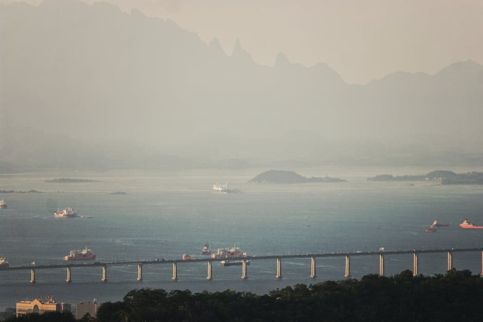 A serene aerial view of a sea bridge connecting coastlines, with ships sailing and mountains in the background.