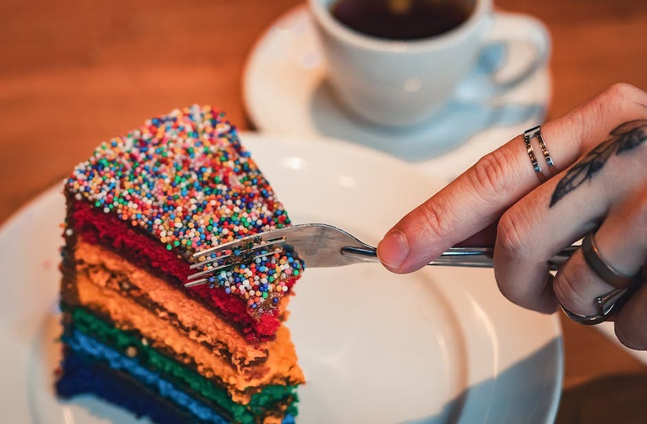 A rainbow layered cake topped with colorful sprinkles is being sliced with a fork in a cozy café setting.