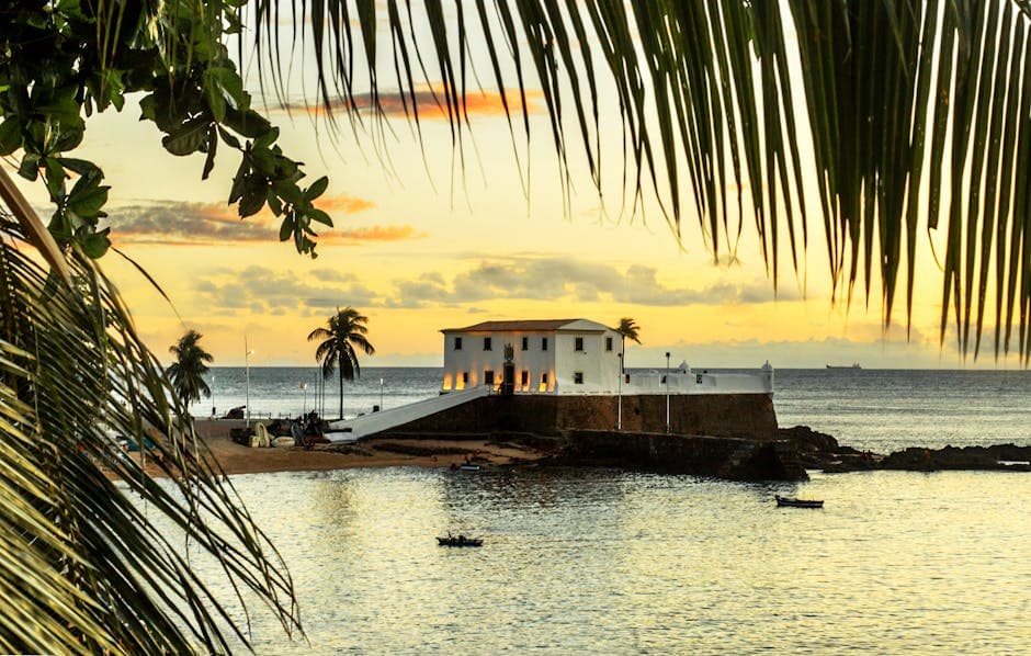 Idyllic sunset view of Forte de Santo Antônio da Barra with palm trees framing the seascape in Salvador, Brazil.