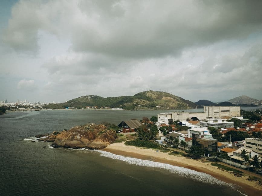 A picturesque aerial view of a beach and urban area in ES, Brazil with lush hills and cloudy skies.