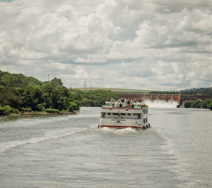 A passenger boat cruises a lush river near a hydroelectric dam on a cloudy day.