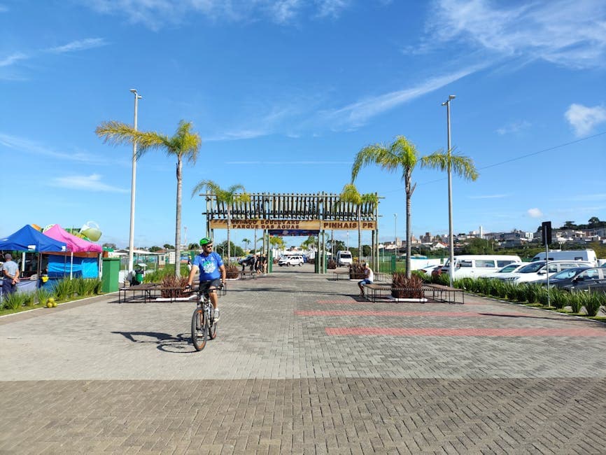 Person biking through a park entrance with clear blue skies and palm trees in Pinhais, Brazil.