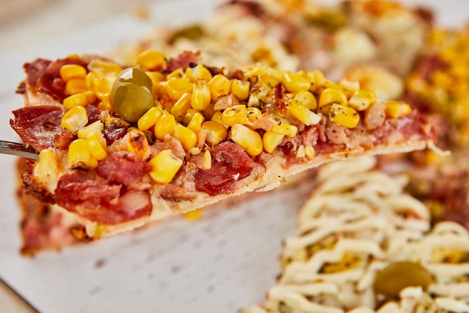 Close-up of a savory pizza slice topped with corn, bacon, and an olive, perfect for food photography.