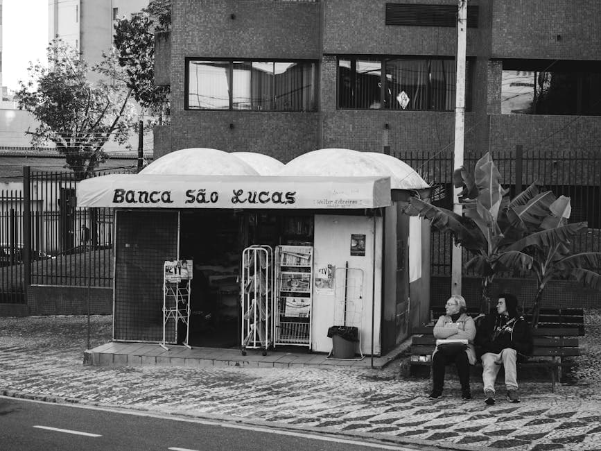 Black and white photo of a newspaper stand with people sitting on a bench.