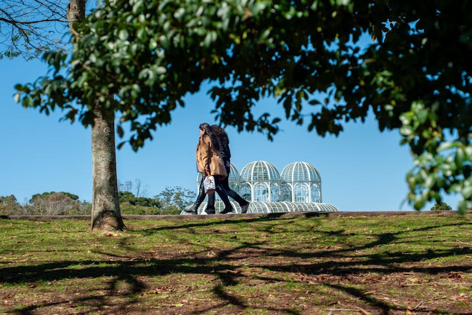 Two women walking outdoors near a botanical garden's glass dome structure, surrounded by trees.