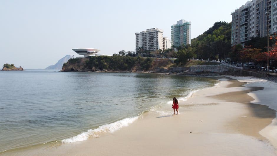 A woman in red strolls on a beach in Niterói, Brazil, with the Museum of Contemporary Art visible in the background.