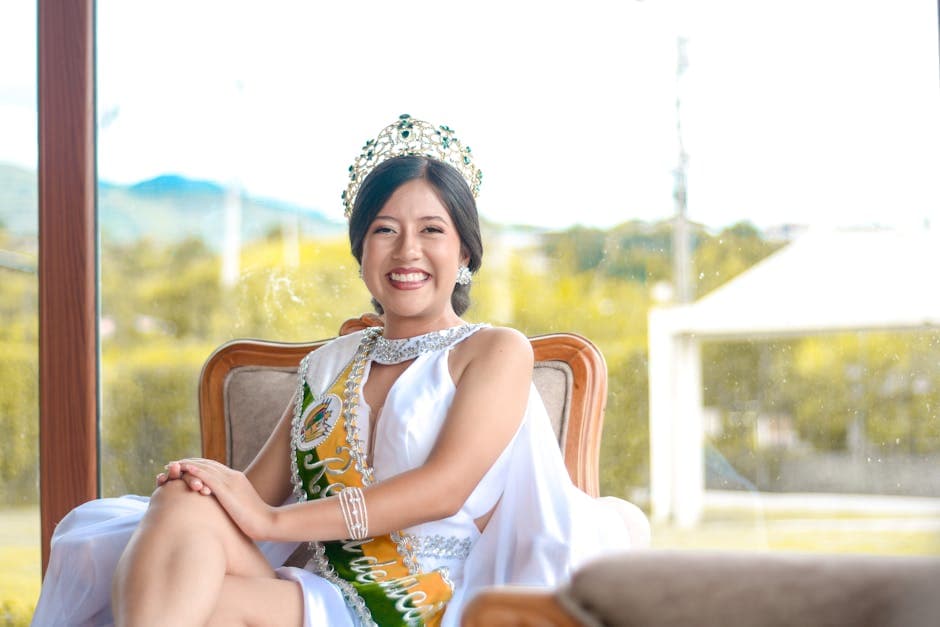 A joyful beauty queen seated elegantly indoors, adorned with a crown and sash, symbolizing grace and celebration.
