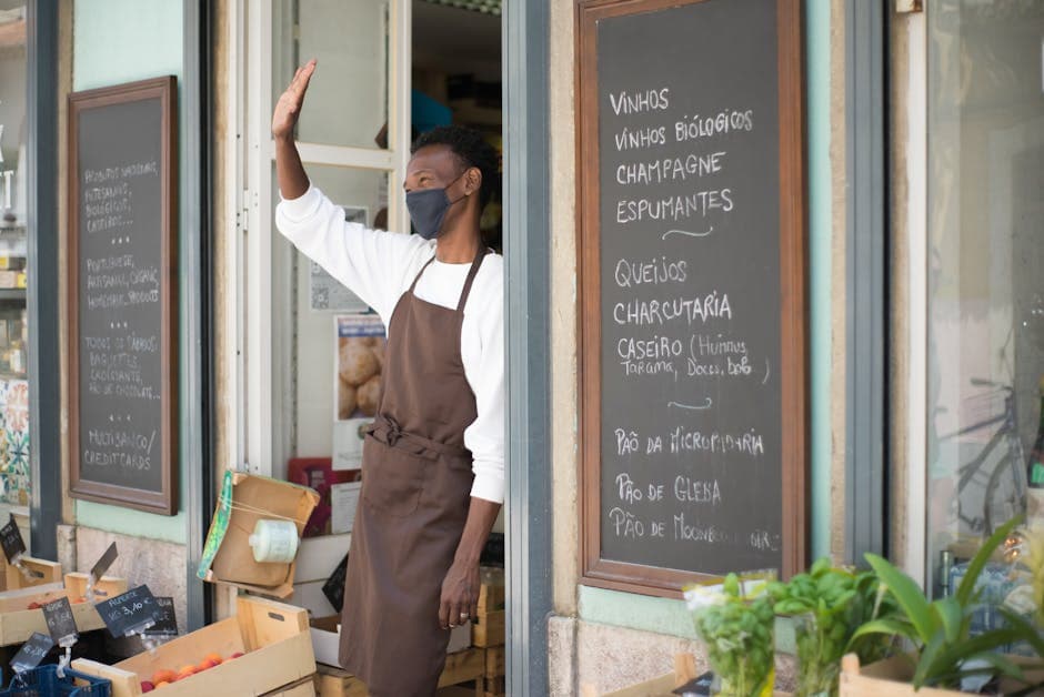 A grocery store clerk wearing an apron and face mask warmly waves from the storefront during pandemic times.