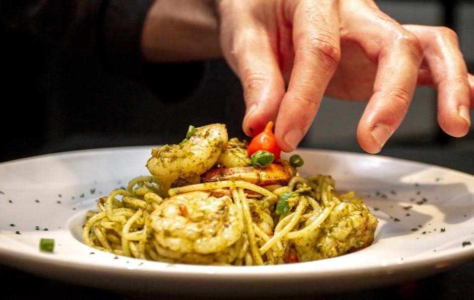 Chef adding final garnish to a plate of shrimp pasta with pesto in a restaurant setting.