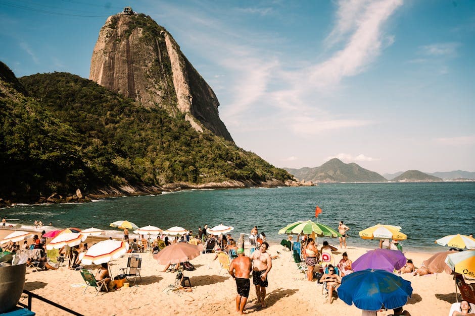 Crowded beach scene under Sugarloaf Mountain in Rio, with vibrant umbrellas and lively atmosphere.