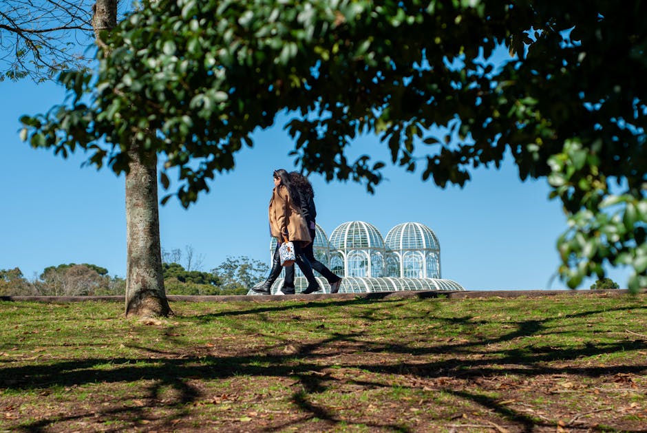 Two women walking outdoors near a botanical garden's glass dome structure, surrounded by trees.