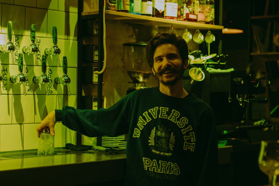 A smiling bartender in a cozy bar wearing a University Paris sweatshirt.